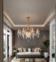An ornate multi-arm chandelier with a gold-toned metal frame and clear crystal droplets, hanging in a modern living room with a gray sofa and dark gray paneled walls.