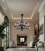 A large multi-light crystal chandelier with black metal arms hangs in a grand, elegant living room with high ceilings, recessed lighting, and modern furniture.