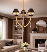 A modern 3-light chandelier with black drum shades, a curved gold-toned frame, and hanging clear crystal pendants, shown suspended in a styled living room with a fireplace, sofa, and mirror.