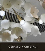White ceramic calla lily pendant lights with clear crystal accents, shown hanging from a ceiling with a dark slatted vent in the background. A dark banner at the bottom reads 'CERAMIC + CRYSTAL'.