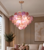 Pink shell disc chandelier suspended in an elegant living room with neutral furnishings, beige wall panelling, and warm ambient lighting.