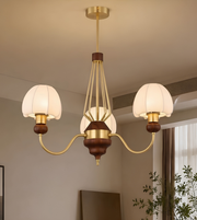 A three-light chandelier with warm brass-finished metal arms, dark brown wooden accents, and white drum-shaped glass shades, shown hanging in a modern interior room.