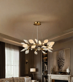 A modern leaf-shaped chandelier with white glass petals and a gold metal frame hangs from the ceiling in a styled living room with dark wood paneling and a fireplace.