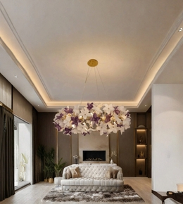 A large modern chandelier with white and purple artificial flowers and gold accents, hanging in a contemporary living room with a tufted sofa and warm ambient lighting.