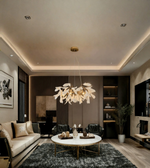 A modern living room interior featuring a large, sculptural leaf-like chandelier with warm white leaves and gold-toned metal accents hanging from the center of a tiered ceiling. The room is styled with a cream sofa, a round white coffee table with gold legs, a dark gray shag rug, and dark wood paneling in the background.