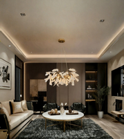 A modern living room interior featuring a large, sculptural leaf-like chandelier with warm white leaves and gold-toned metal accents hanging from the center of a tiered ceiling. The room is styled with a cream sofa, a round white coffee table with gold legs, a dark gray shag rug, and dark wood paneling in the background.