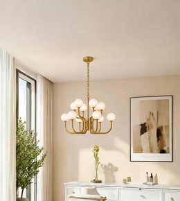 A modern brass chandelier with nine frosted glass globe shades hangs in a bright, contemporary room with beige walls, a white vanity, and a window with sheer curtains.
