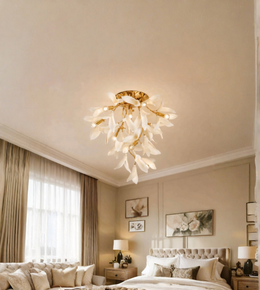 A modern chandelier with white petal-like glass shades and a gold metal frame, hanging from the ceiling in a styled bedroom with beige walls, a bed, and curtains.