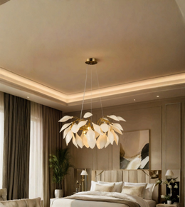 A modern pendant chandelier with a cluster of white leaf-shaped petals and gold-toned metal accents, hanging in a styled bedroom with warm ambient lighting.