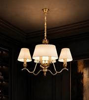A three-light brass chandelier with white drum shades hanging from a twisted chain in a dark, elegant room.