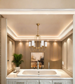 A modern luxury bathroom interior featuring a gold multi-light chandelier with white cylindrical shades hanging over a white double vanity and integrated bathtub. The background consists of warm beige paneled walls, recessed lighting, and a framed abstract painting.
