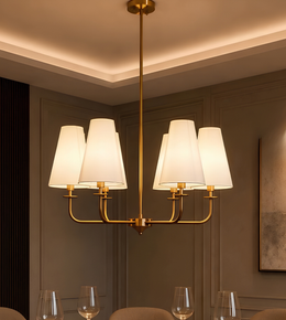A modern six-light brass chandelier with white tapered fabric shades, hanging in a warm, elegantly lit dining room.