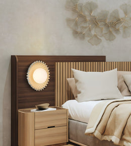 A modern bedroom scene featuring a wooden headboard with an integrated circular sunburst wall lamp, a matching light wood nightstand, and neutral-toned bedding against a textured beige wall.