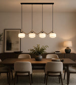 A modern multi-light linear pendant chandelier with four glowing white cylindrical shades and gold-toned geometric accents, hanging over a dining table in a warm, contemporary dining room.