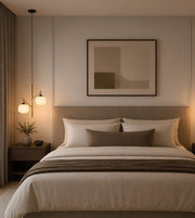 A modern bedroom featuring a neatly made bed with white and taupe bedding, a matching headboard, a framed abstract print on the wall, and warm pendant lights on a nightstand.