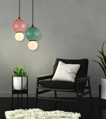 A modern living room scene featuring two hanging pendant lights with pink, green, and white globe shades, a black armchair with a white pillow, and small potted plants against a gray wall.