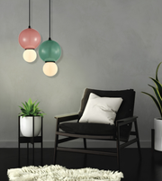 A modern living room scene featuring two hanging pendant lights with pink, green, and white globe shades, a black armchair with a white pillow, and small potted plants against a gray wall.