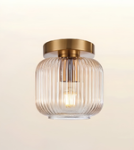 A modern semi-flush mount ceiling light fixture featuring a ribbed clear glass shade and a brushed brass gold ceiling canopy, shown against a warm off-white background.